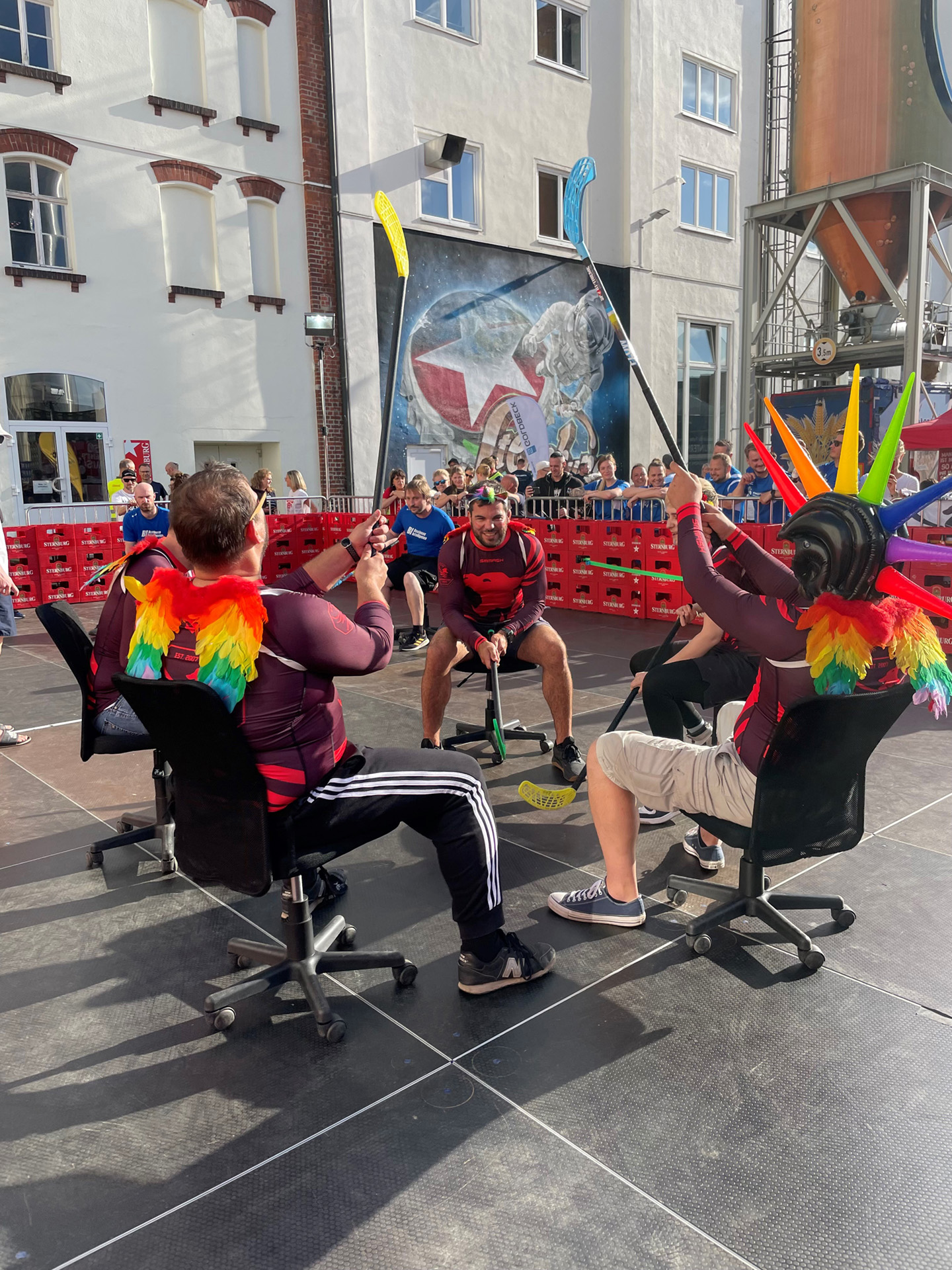 Drei Personen sitzen auf Bürostühlen und ziehen an Seilen bei einem Event.