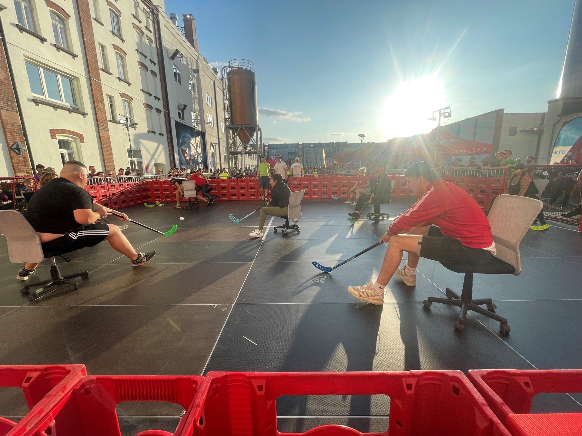 Zwei Personen sitzen auf Bürostühlen und spielen Hockey auf einem Platz in einer städtischen Umgebung.