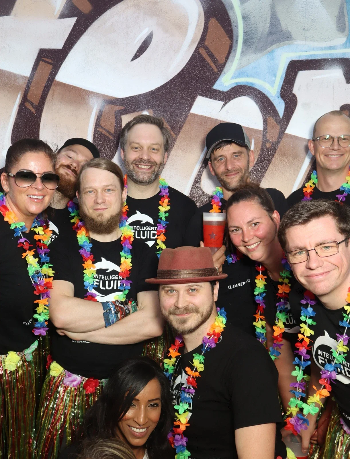 Eine Gruppe von Menschen trägt schwarze T-Shirts und bunte Blumenketten. Der Hintergrund zeigt eine Graffiti-Wand.