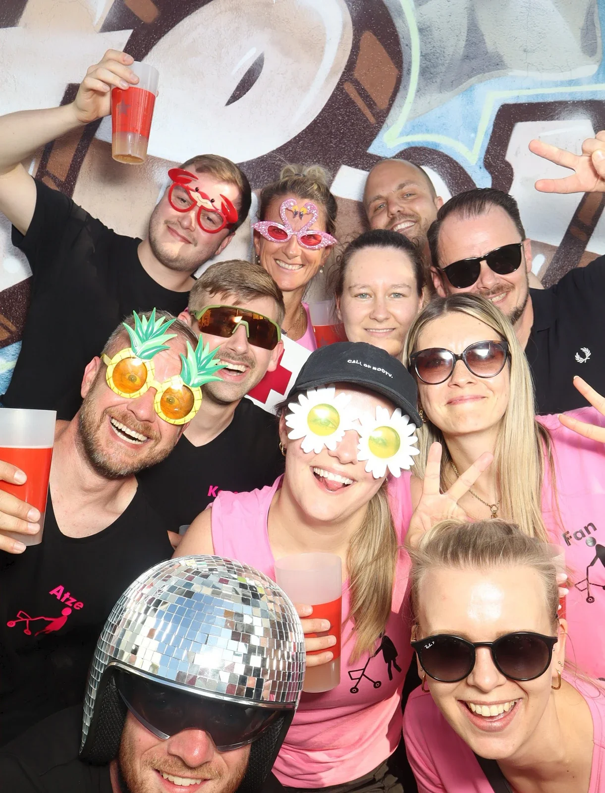 Elf Personen tragen bunte Brillen und halten Getränke in der Hand. Sie stehen vor einer Wand mit Graffiti.