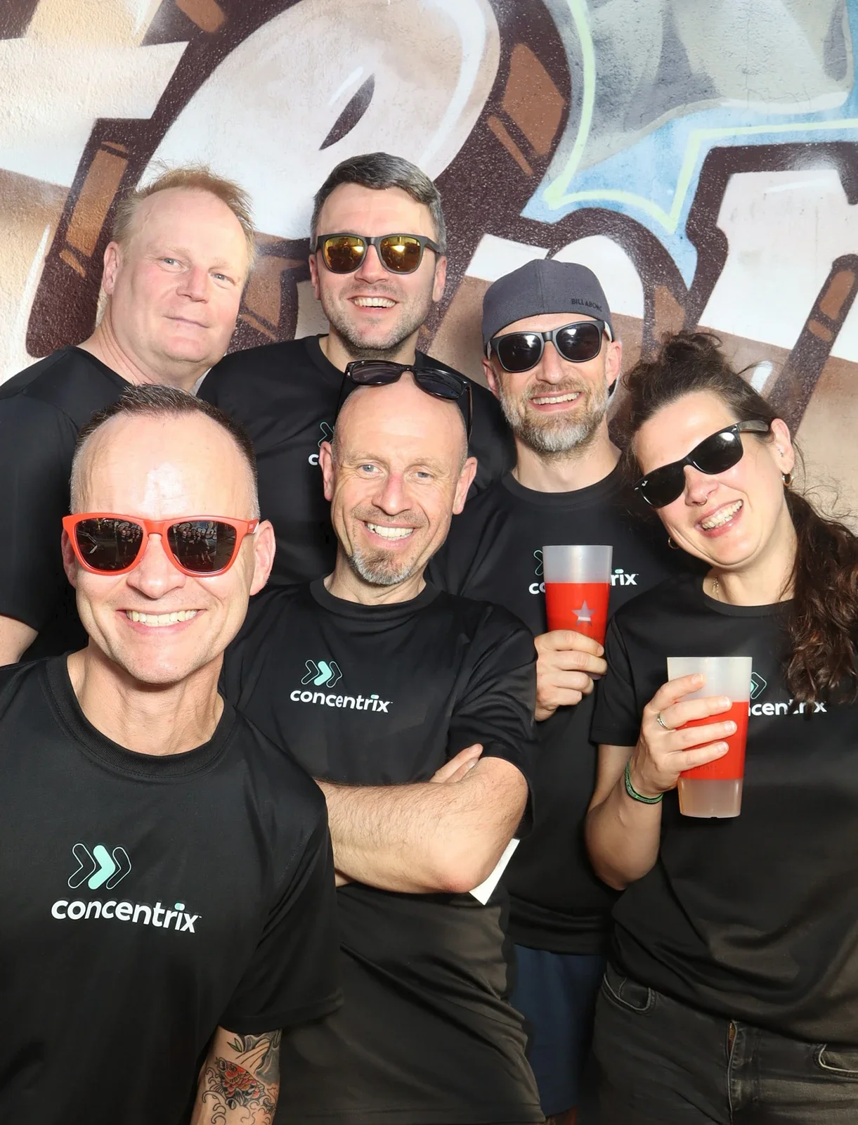 Sechs Personen in schwarzen Trikots mit Sonnenbrillen halten rote Getränke. Im Hintergrund ist eine Graffiti-Wand.