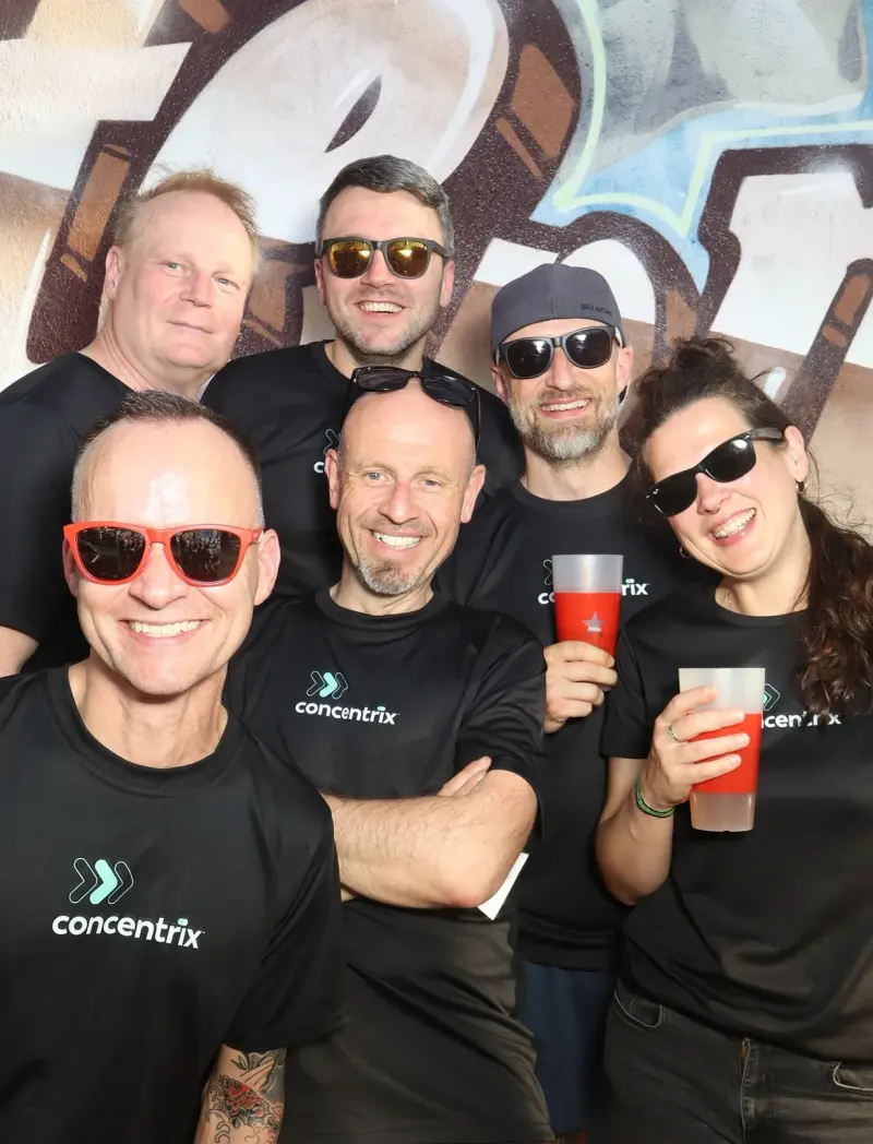 Sechs Personen in schwarzen Trikots mit Sonnenbrillen halten rote Getränke. Im Hintergrund ist eine Graffiti-Wand.