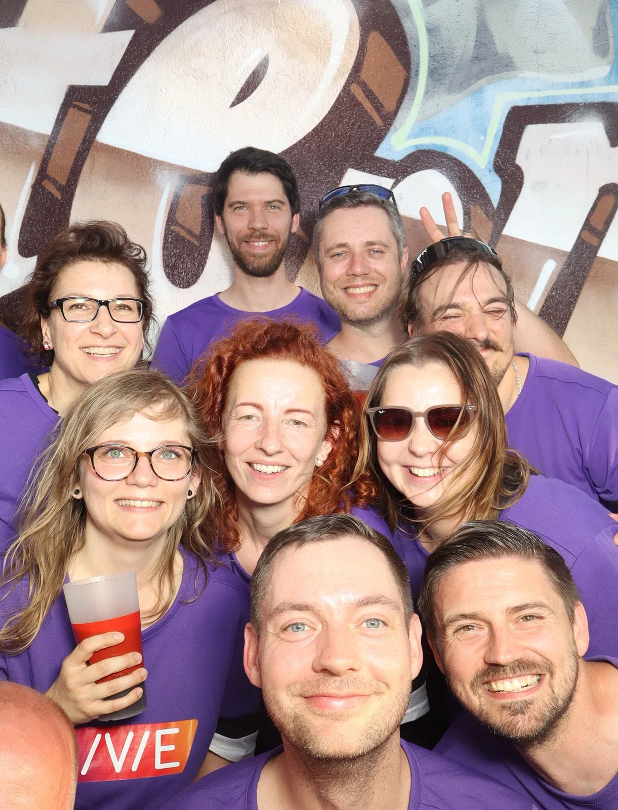 Zehn Personen in lila Trikots stehen dicht beieinander und lächeln. Der Hintergrund zeigt eine bemalte Wand.