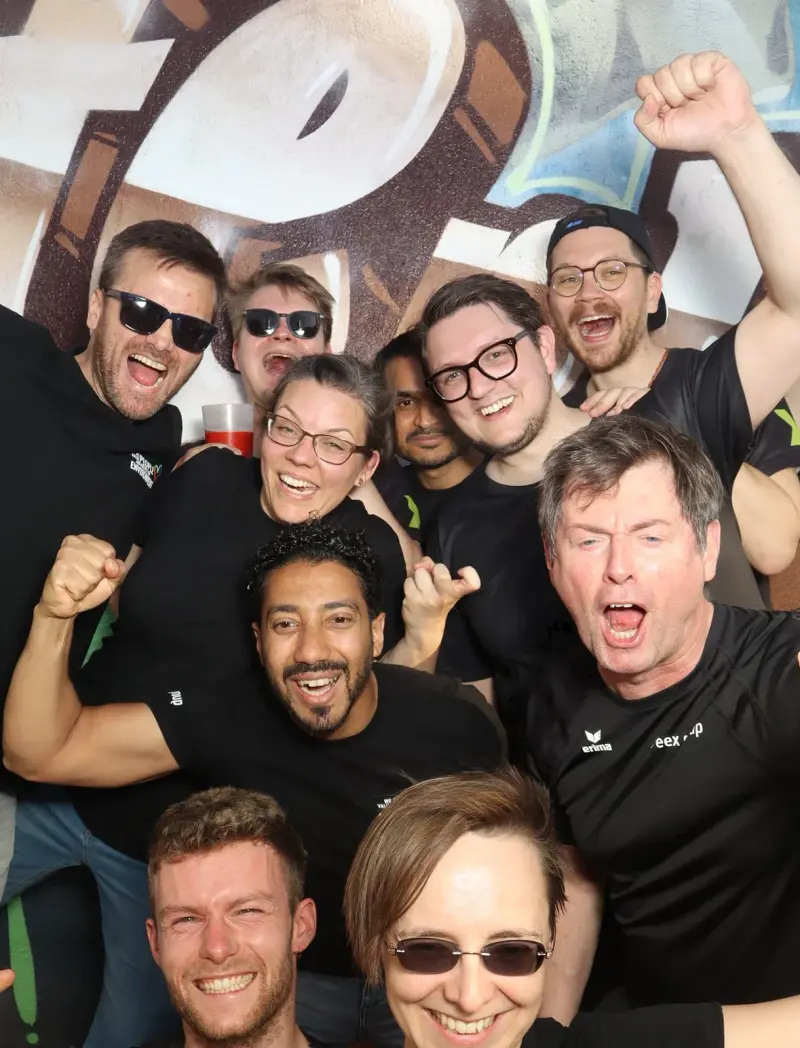 Zehn Personen in schwarzen Shirts posieren lachend und jubelnd. Im Hintergrund ist eine Wand mit Graffiti.