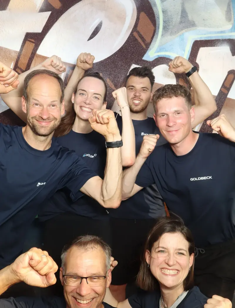 Sieben Personen in dunkelblauen Trikots zeigen Muskelposen. Der Hintergrund ist mit Graffiti bemalt.