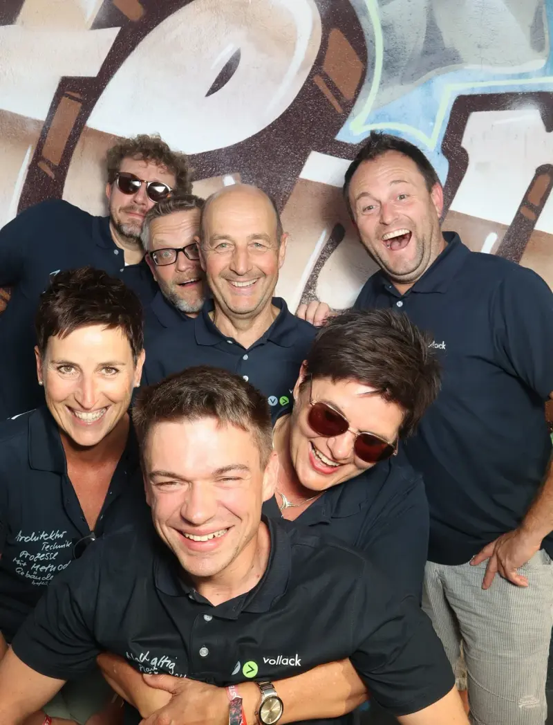 Acht Personen in dunkelblauen Poloshirts stehen dicht beieinander und lächeln. Im Hintergrund eine Graffiti-Wand.