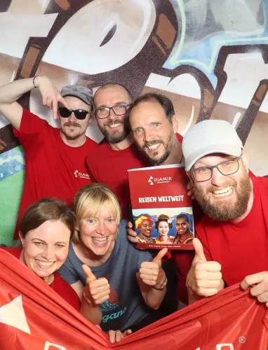 Acht Personen in roten Shirts halten eine rote Fahne und ein Plakat. Alle zeigen Daumen hoch vor Graffiti-Wand.