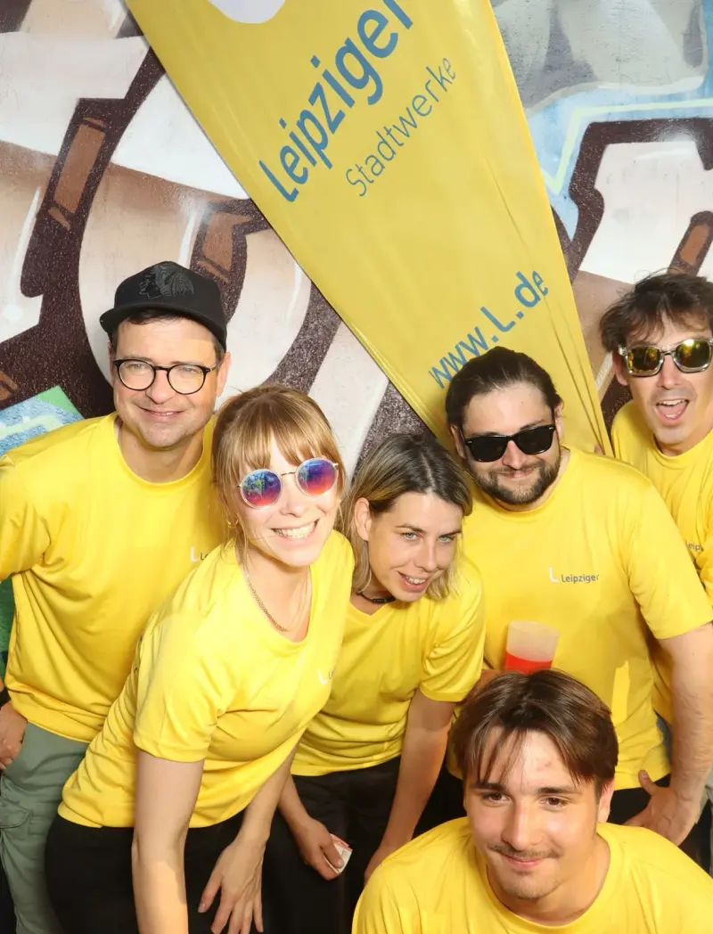 Sechs Personen in gelben Trikots stehen vor einem gelben Banner. Alle tragen Sonnenbrillen oder lächeln in die Kamera.