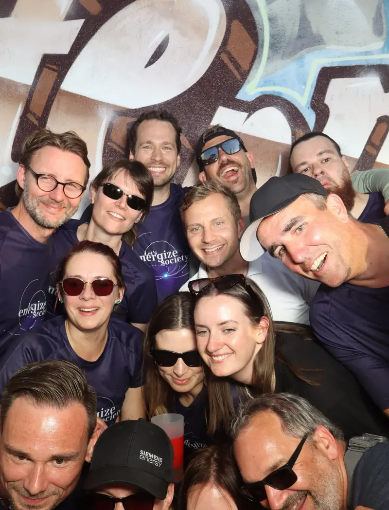 Vierzehn Personen in dunklen Trikots stehen dicht beieinander und lächeln. Im Hintergrund ist eine bemalte Wand.