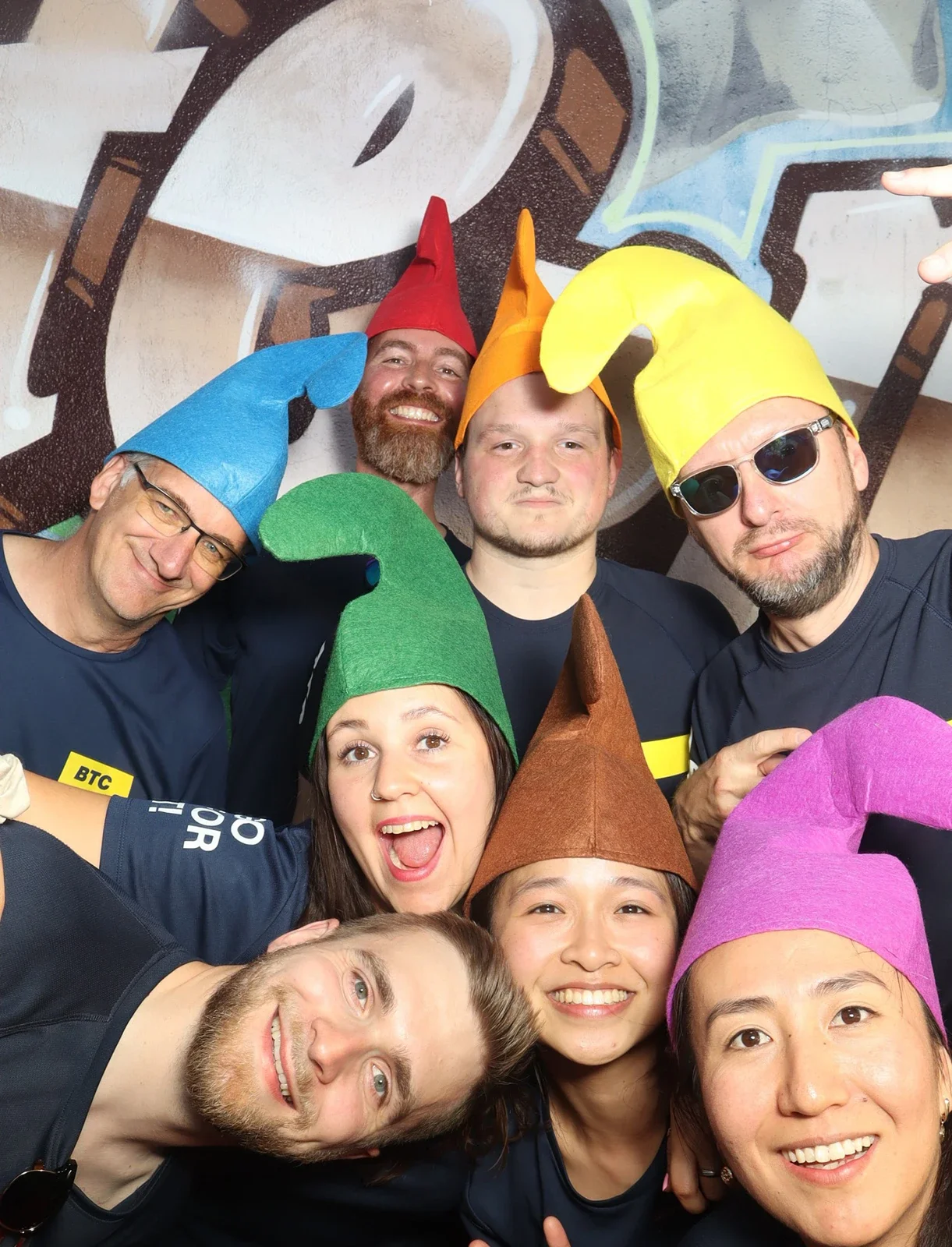 Acht Personen tragen spitze bunte Mützen und dunkelblaue Trikots. Sie stehen vor einer Wand mit Graffiti.