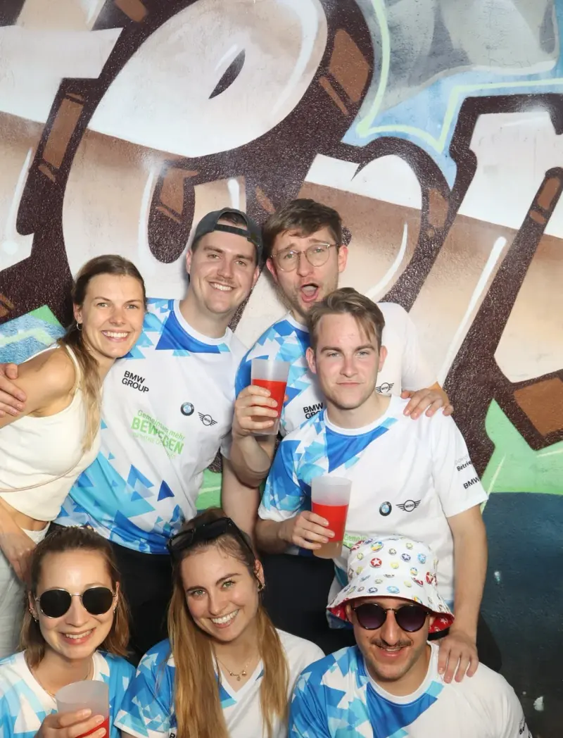 Acht Personen in weiß-blauen Trikots halten rote Getränke in der Hand. Der Hintergrund ist mit Graffiti gestaltet.