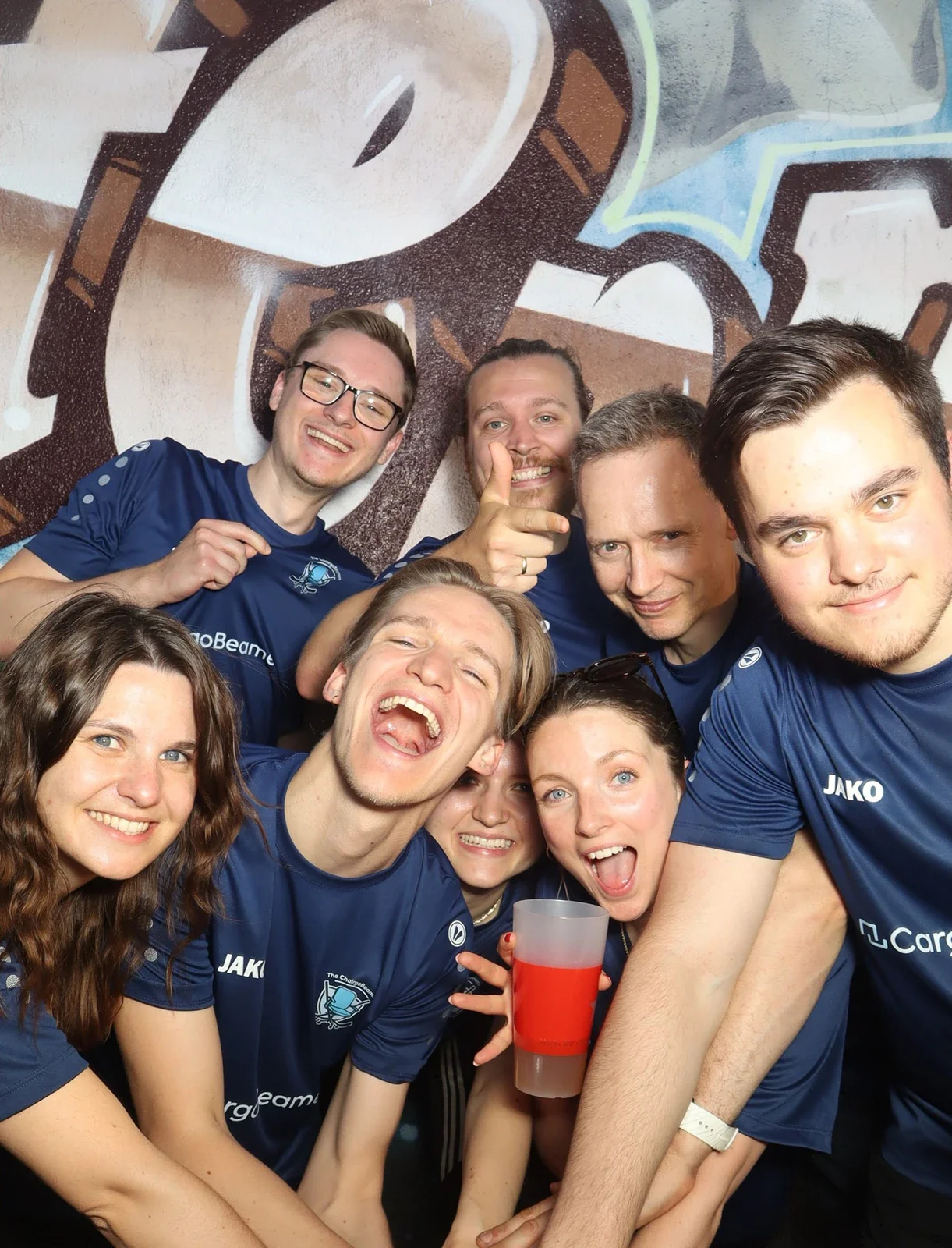 Acht Personen in blauen Trikots stehen eng beieinander. Einige lachen, andere halten ein Getränk in der Hand.