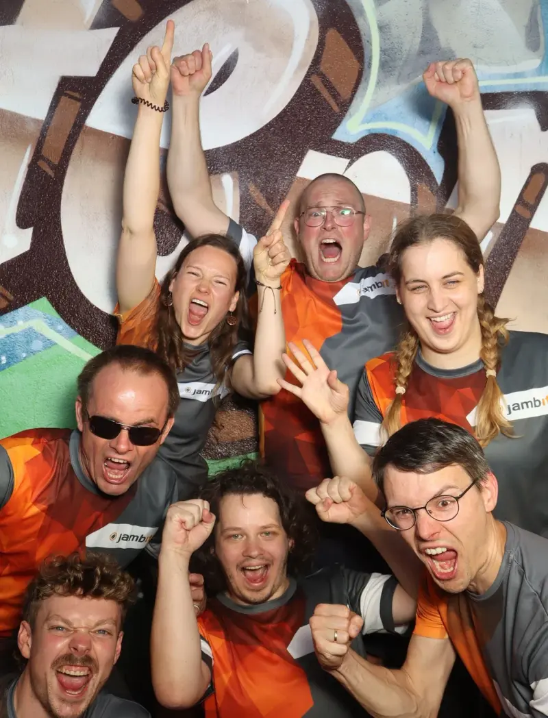 Acht Personen in grau-orangenen Trikots jubeln mit erhobenen Armen. Der Hintergrund zeigt eine bemalte Wand.