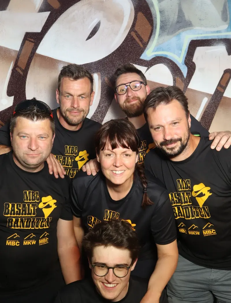 Sieben Personen in schwarzen Shirts mit gelbem Aufdruck stehen dicht beieinander und blicken in die Kamera.