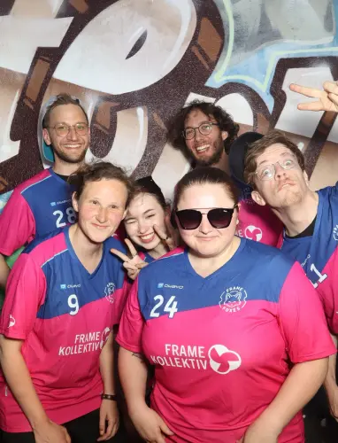 Sieben Personen in pink-blauen Trikots stehen vor einer Graffiti-Wand. Zwei zeigen ein Peace-Zeichen.