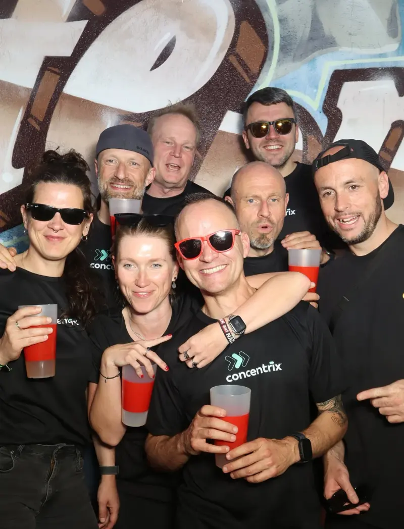 Acht Personen in schwarzen Shirts mit Logo halten rote Getränke. Sie stehen eng beisammen vor einer bemalten Wand.