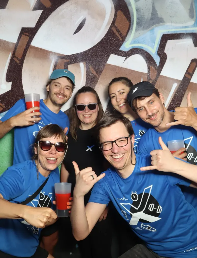 Sechs Personen in blauen Shirts posieren mit Getränken. Einige zeigen Gesten, im Hintergrund ist Graffiti.