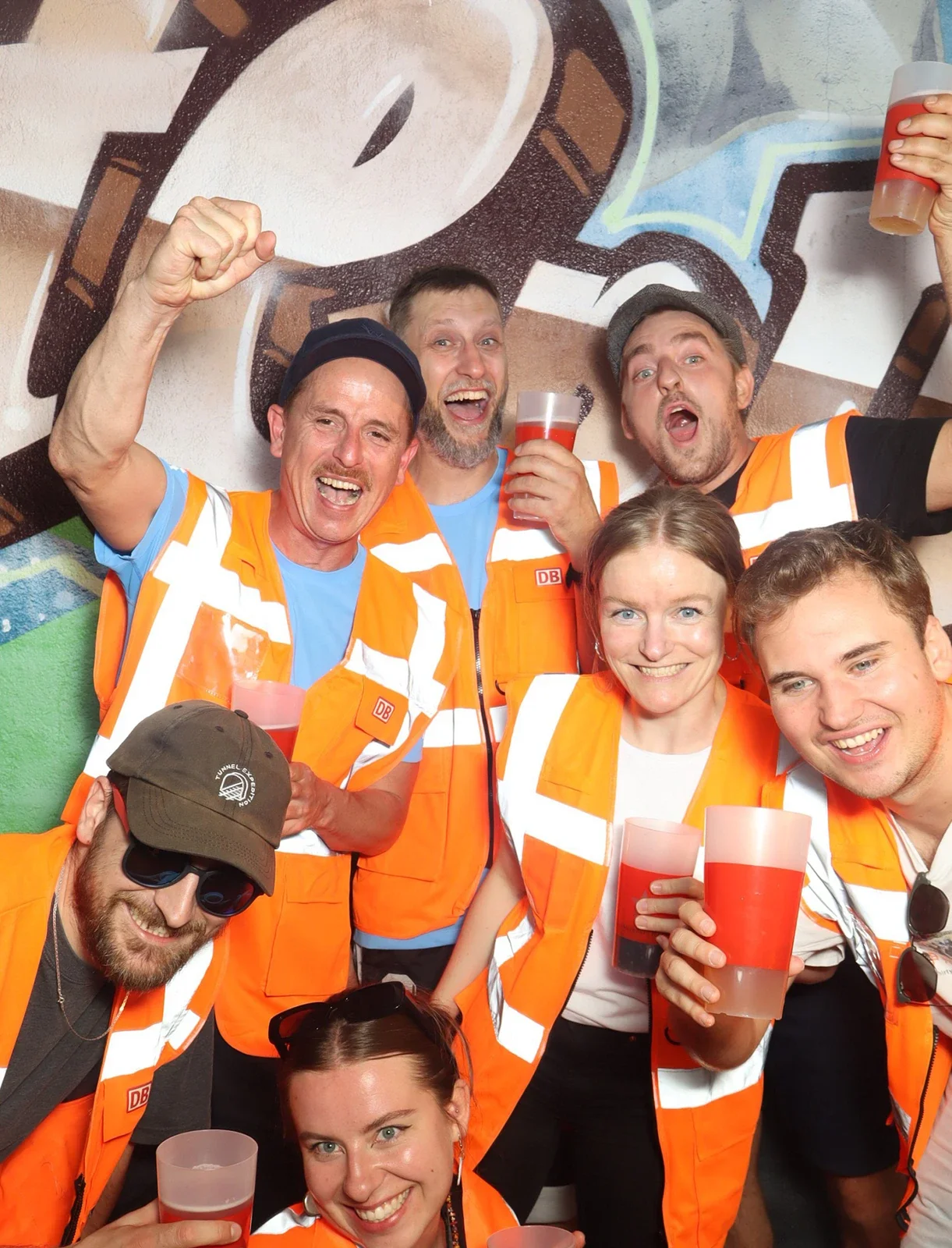 Acht Personen in Warnwesten der Bahn halten rote Getränke. Alle lächeln und stehen dicht vor einer Graffiti-Wand.