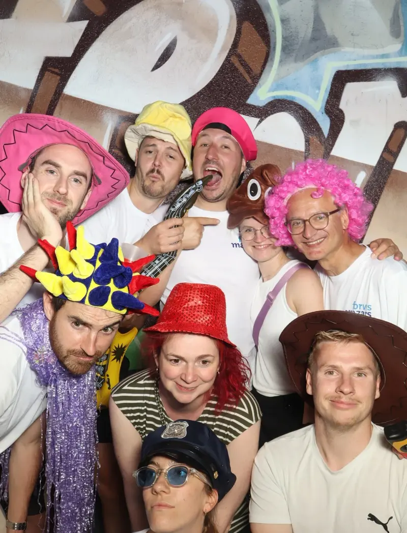 Zwölf Personen mit bunten Hüten und Accessoires stehen zusammen. Der Hintergrund zeigt eine bemalte Wand.
