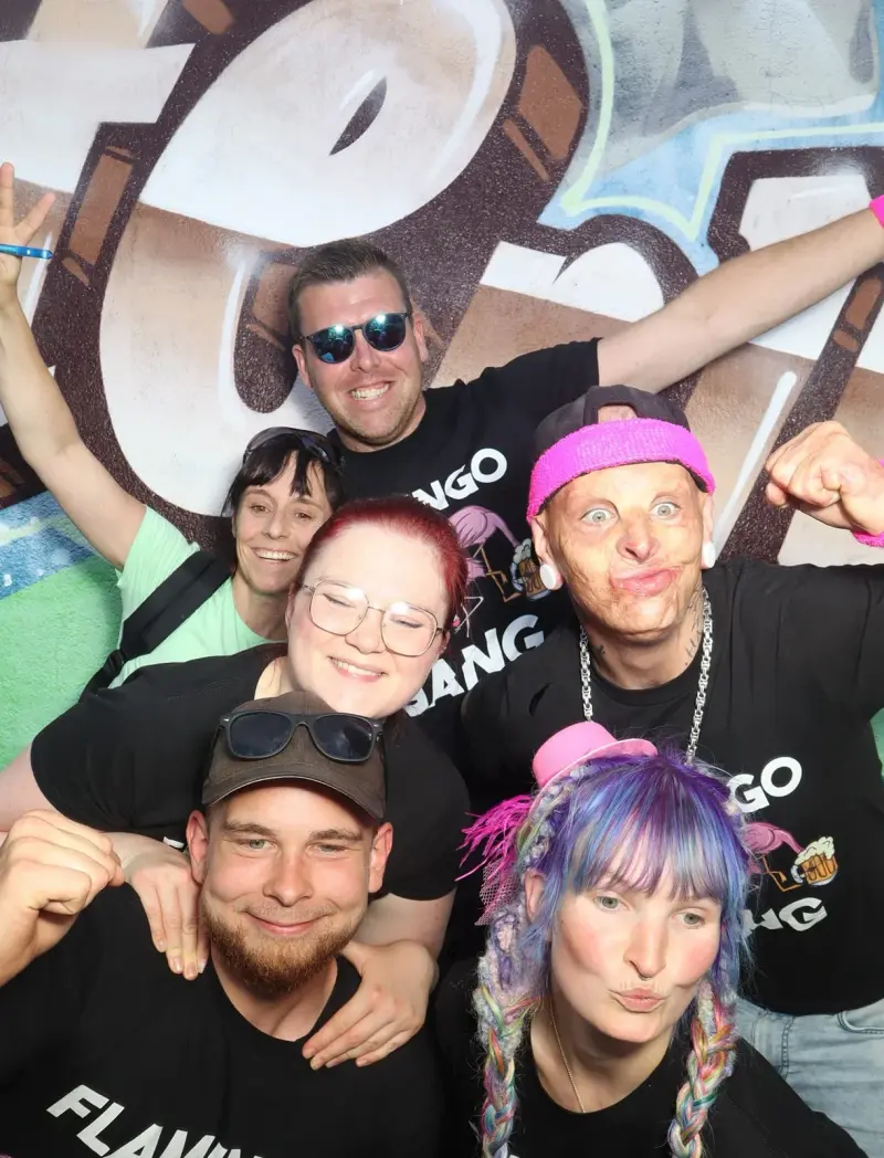 Sechs Personen in schwarzen T-Shirts posieren mit bunten Accessoires. Im Hintergrund ist eine Graffiti-Wand erkennbar.