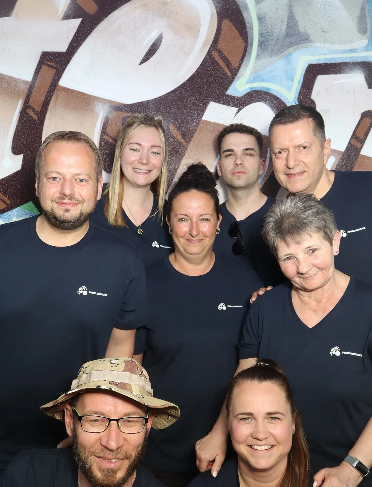 Acht Personen in dunkelblauen T-Shirts stehen eng beieinander. Im Hintergrund ist eine bunte Wand mit Graffiti.