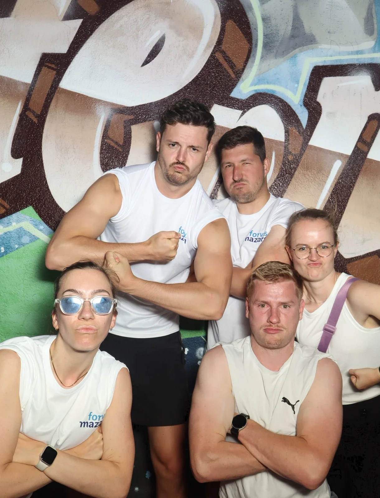 Fünf Personen in weißen Shirts posieren mit verschränkten Armen und ernsten Gesichtern. Der Hintergrund zeigt Graffiti.