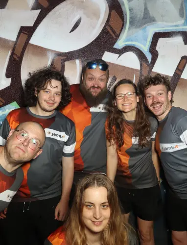 Sieben Personen in grau-orangenen Trikots stehen eng beieinander. Einige lachen, andere schauen ernst.