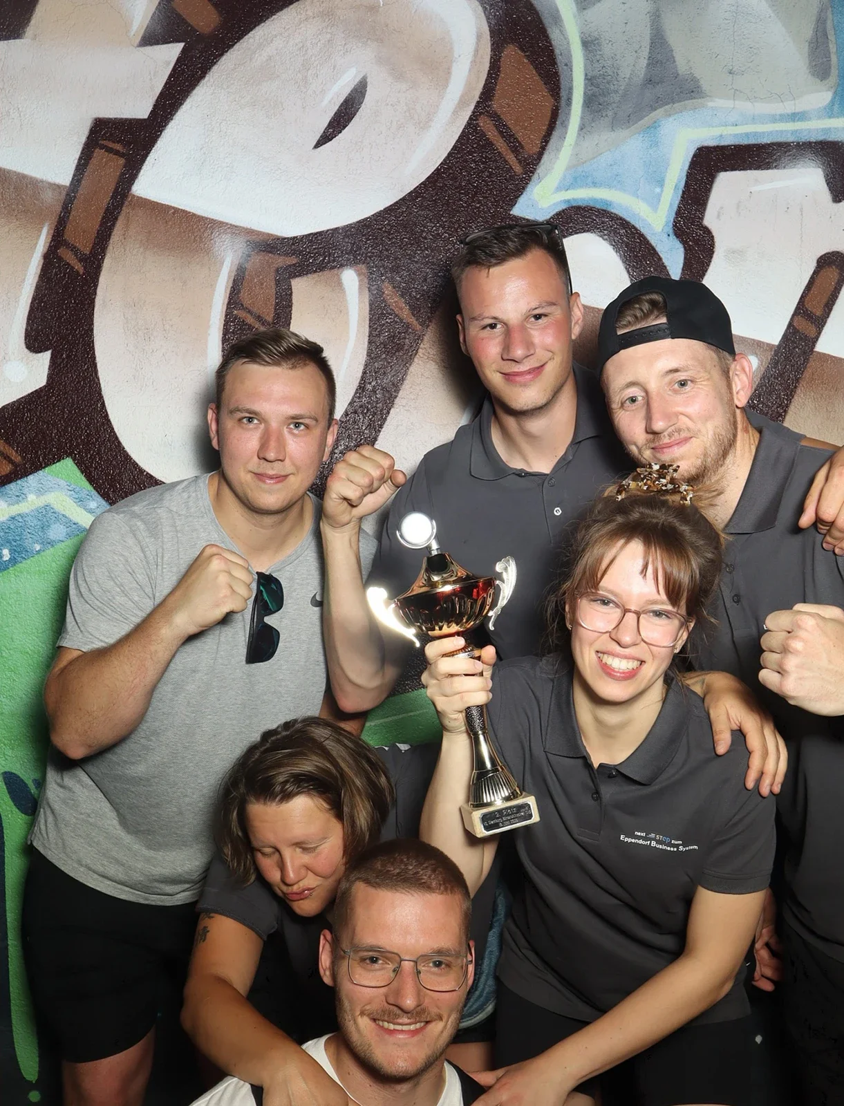 Sieben Personen posieren mit Pokal in der Mitte. Alle tragen graue Shirts, im Hintergrund eine bunte Wand.