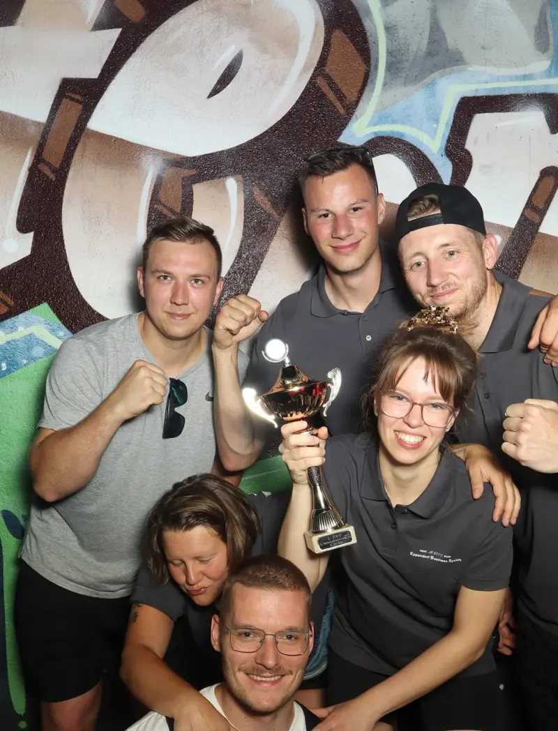 Sieben Personen posieren mit Pokal in der Mitte. Alle tragen graue Shirts, im Hintergrund eine bunte Wand.