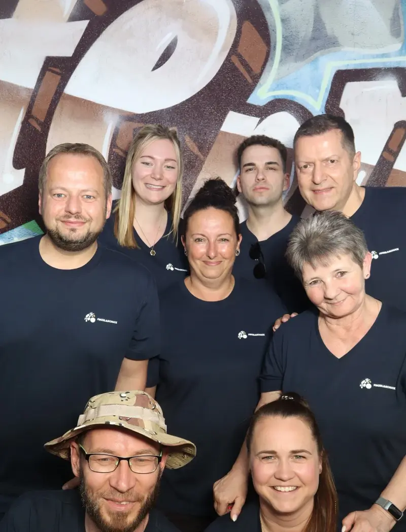 Acht Personen in dunkelblauen T-Shirts stehen eng beieinander. Im Hintergrund ist eine bunte Wand mit Graffiti.