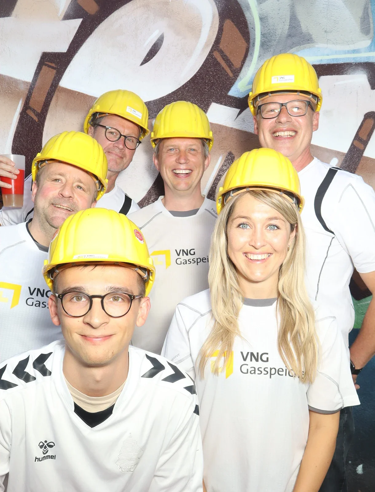 Sechs Personen tragen gelbe Schutzhelme und weiße Trikots mit Firmenlogo. Im Hintergrund ist eine bemalte Wand.