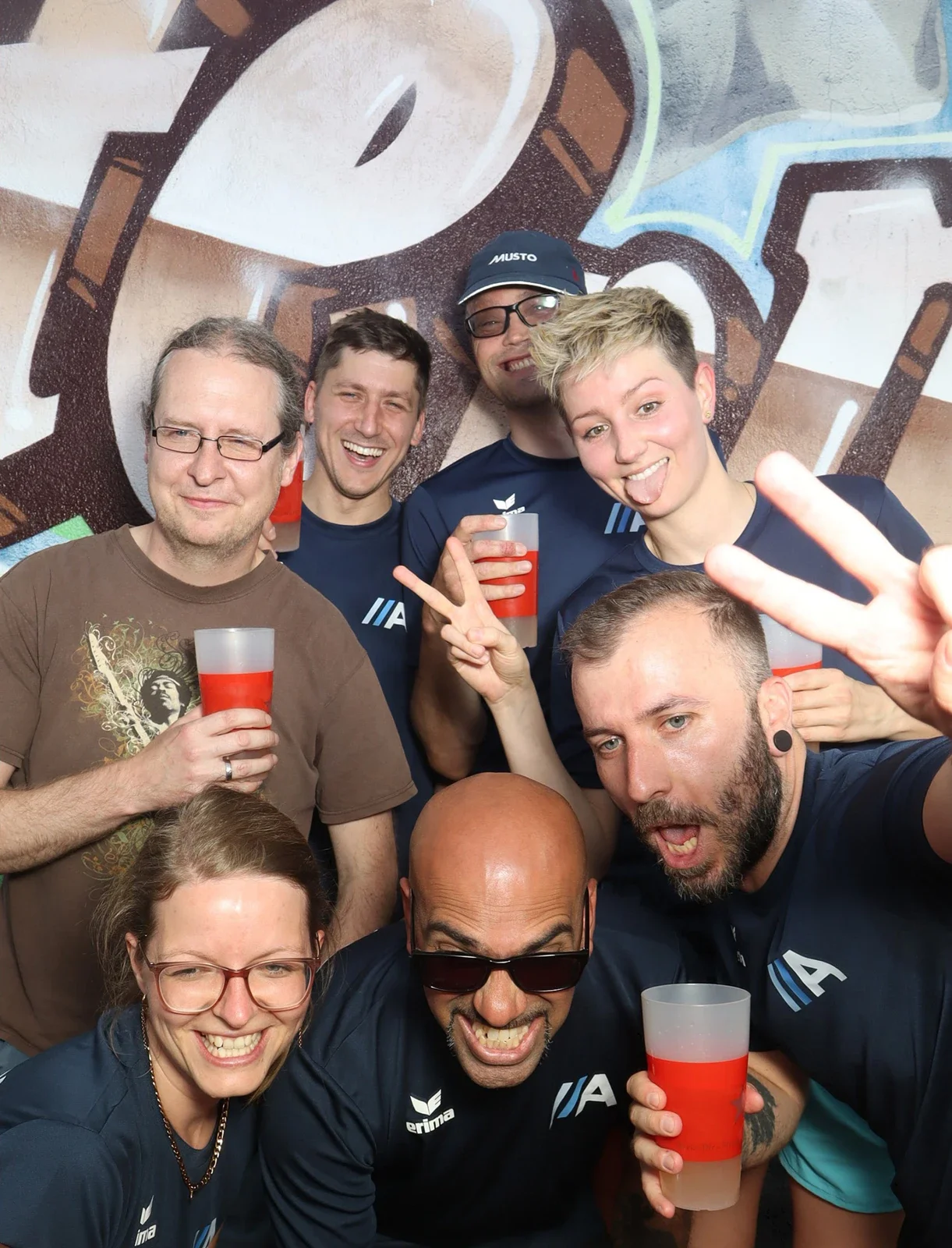 Acht Personen posieren mit roten Getränken in der Hand. Sie tragen dunkle Shirts und stehen vor einer Graffiti-Wand.