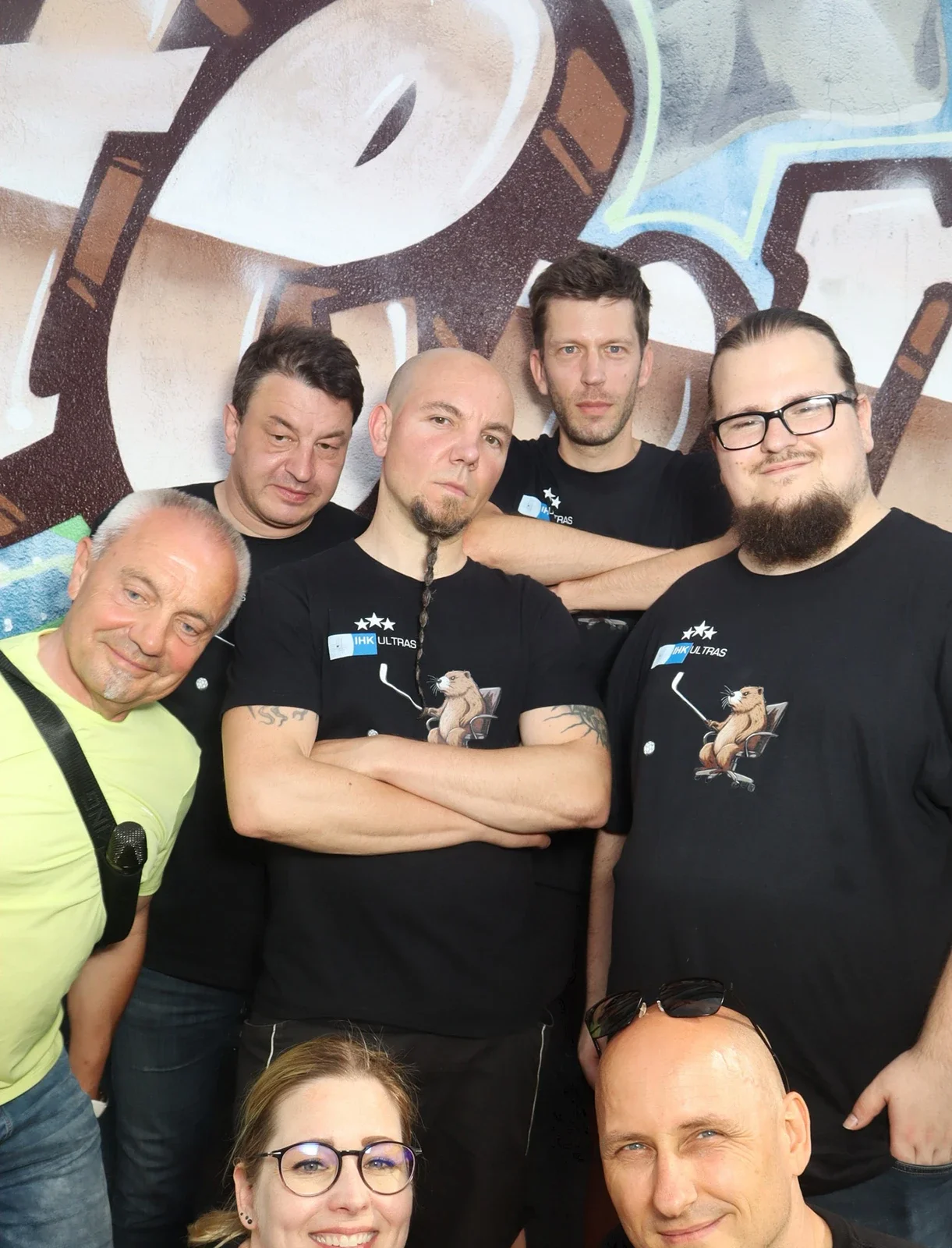 Acht Personen stehen in schwarzer Kleidung vor einer Graffiti-Wand. Einige haben verschränkte Arme oder lächeln in die Kamera.