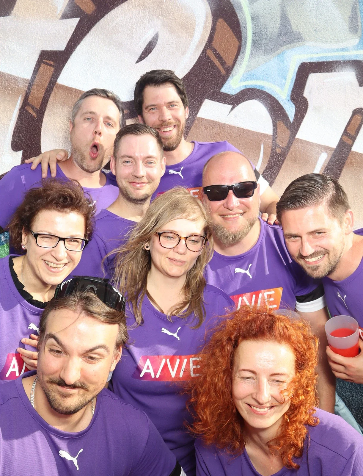Neun Personen in lila Trikots stehen eng beieinander. Einige lächeln, andere machen Grimassen vor einer bemalten Wand.