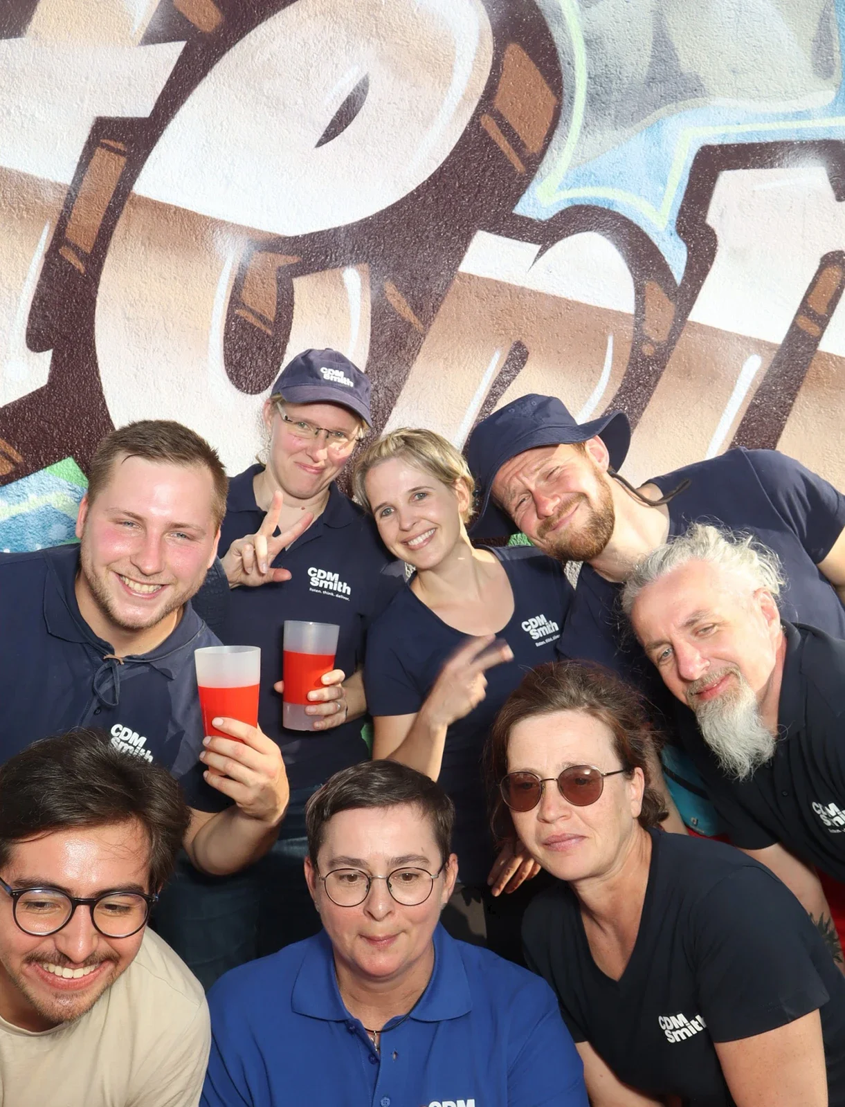 Acht Personen in dunkelblauer Kleidung posieren vor einer Graffiti-Wand. Einige halten Becher mit rotem Getränk.