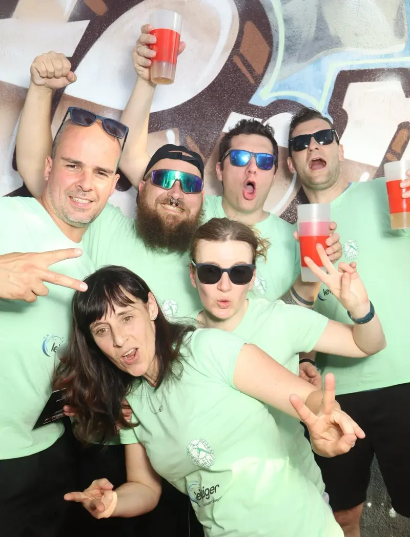 Sieben Personen in hellgrünen Shirts posieren gestikulierend mit roten Getränken. Im Hintergrund eine bemalte Wand.