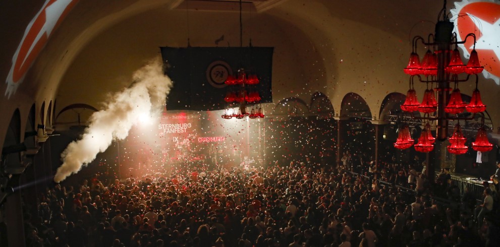 Eine große Menschenmenge genießt ein Konzert in einer beleuchteten Halle. Auf der Bühne wird Konfetti in die Luft geblasen.