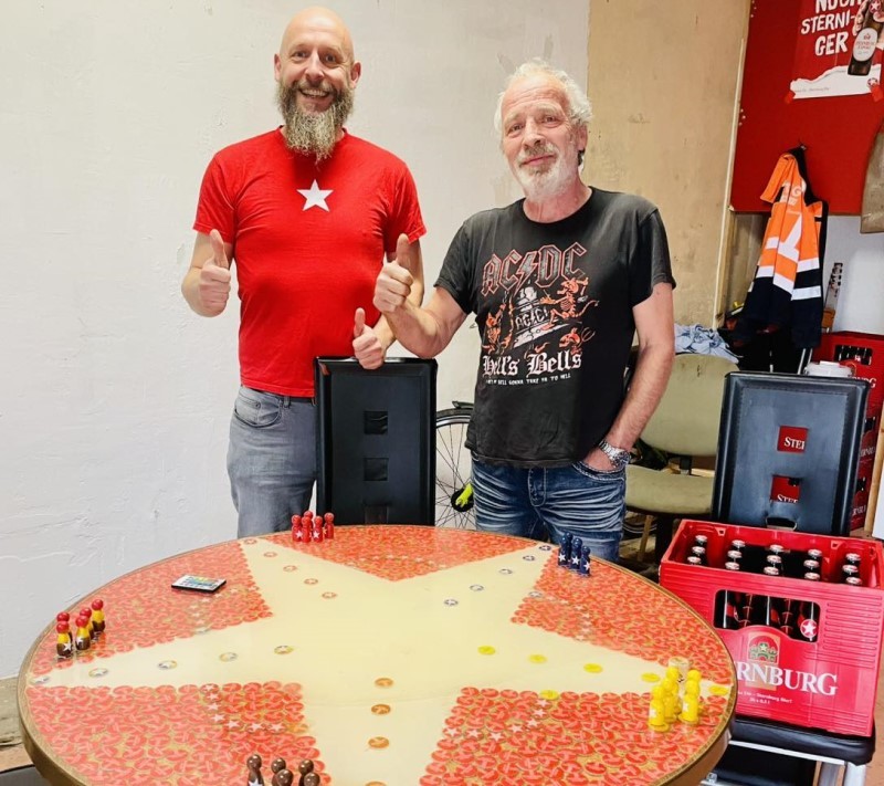 Zwei Männer stehen lächelnd an einem runden Tisch bestehend aus jeder Menge Sternburg-Kronkorken. Der Tisch ist ein Mensch-Ärgere-Dich-Nicht-Spieltisch.