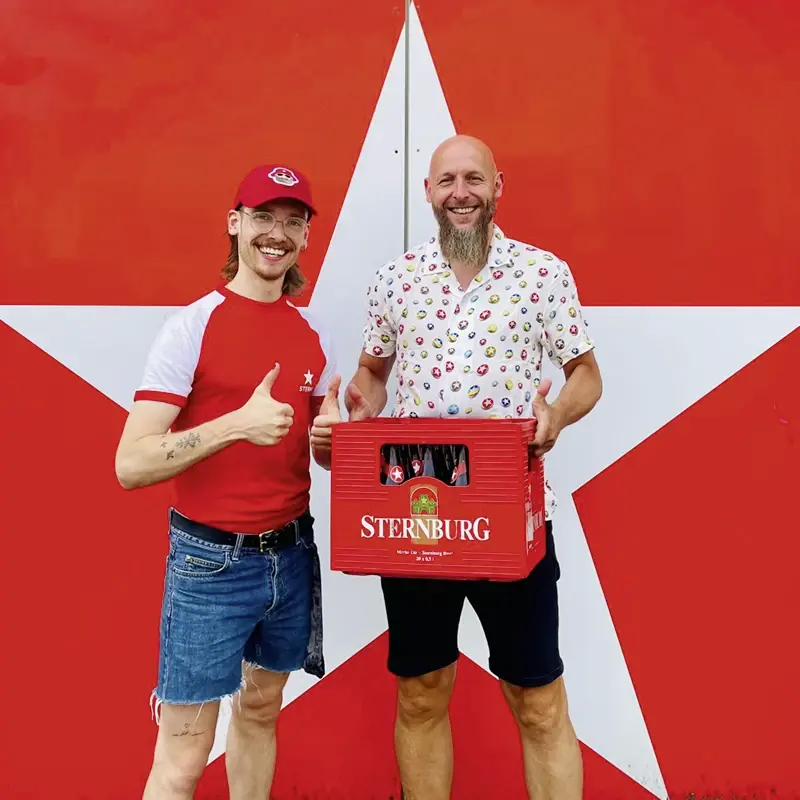 Zwei Männer stehen vor einer roten Wand mit weißem Stern, einer hält einen roten Sternburg-Bierkasten in der Hand.