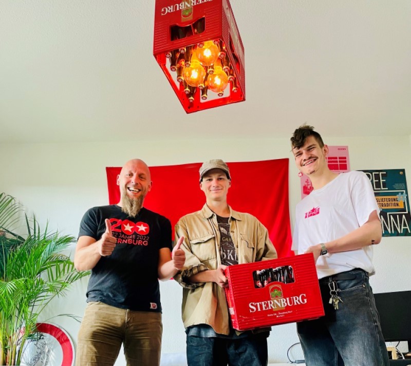Drei lächelnde Männer stehen in einem Wohnzimmer. Zwei davon halten den Sternburg Bierkasten des Monats hoch. An der Decke hängt eine Lampe im Bierkasten-Design.