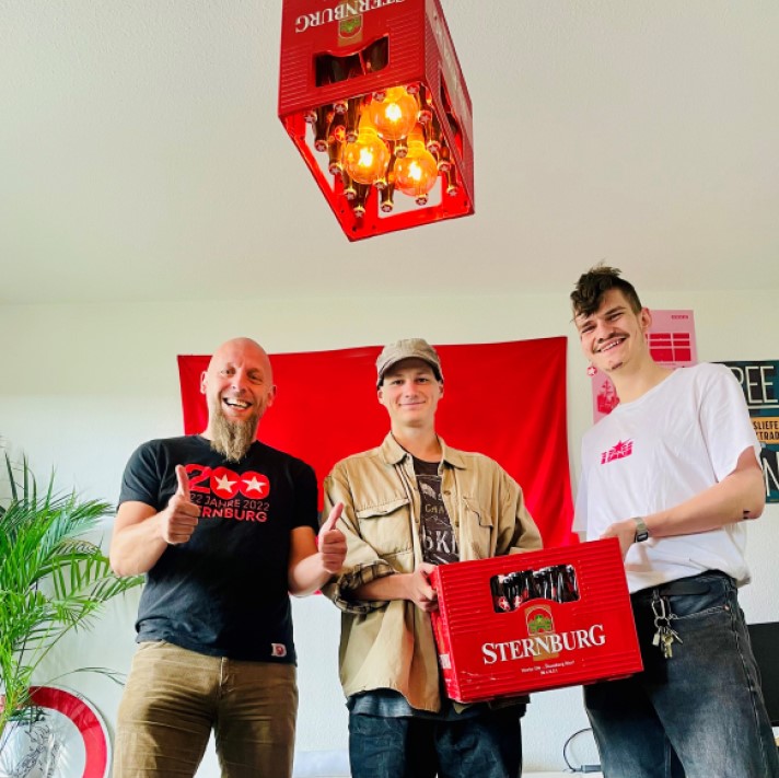 Drei lächelnde Männer stehen in einem Wohnzimmer. Zwei davon halten den Sternburg Bierkasten des Monats hoch. An der Decke hängt eine Lampe im Bierkasten-Design.