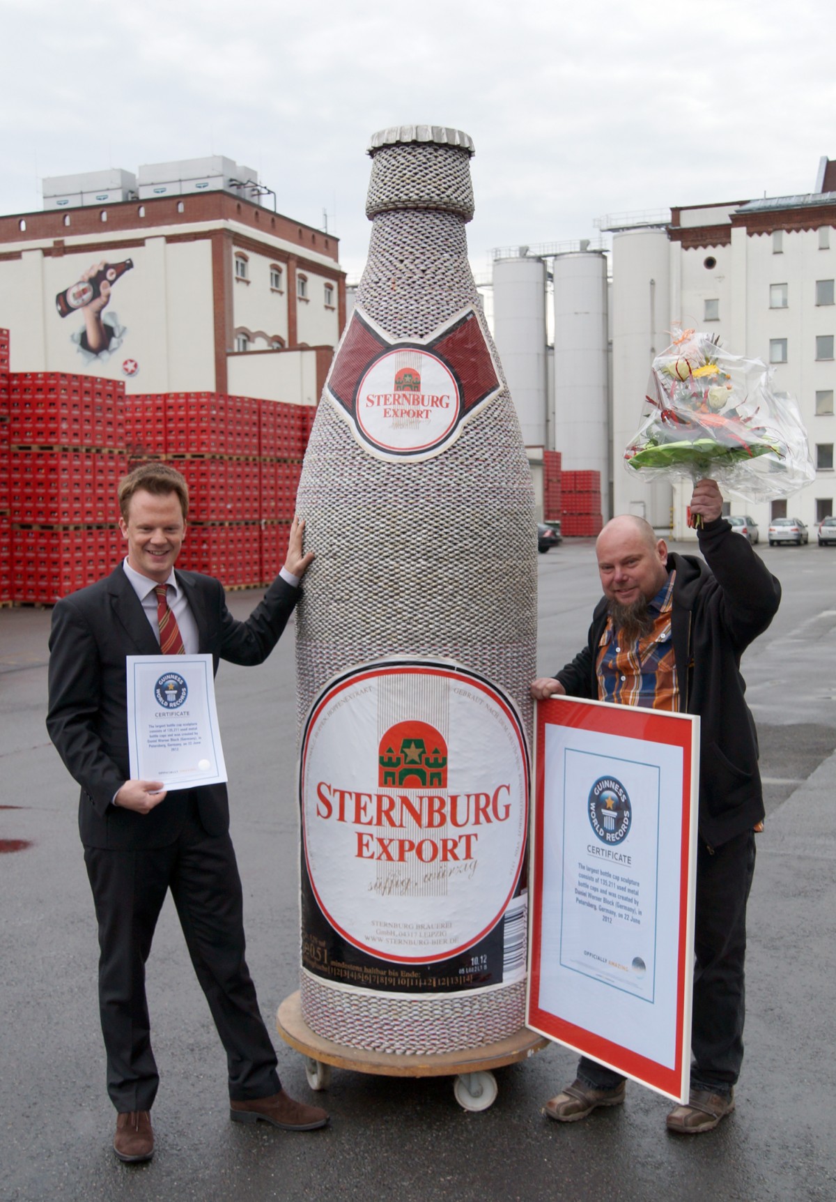 Zwei Männer stehen neben einer großen Flaschenattrappe und halten Zertifikate.