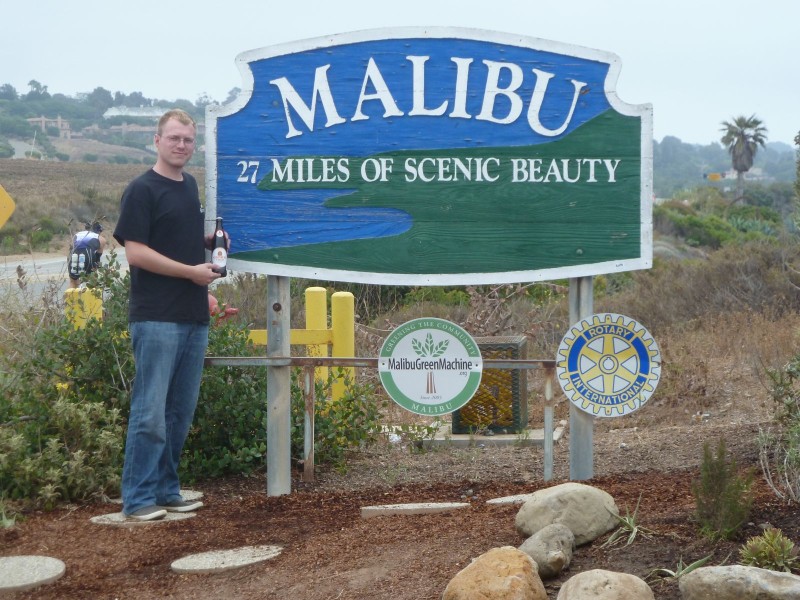 Eine Person steht neben einem Schild mit der Aufschrift Malibu und hält eine Flasche Sternburg in der Hand.