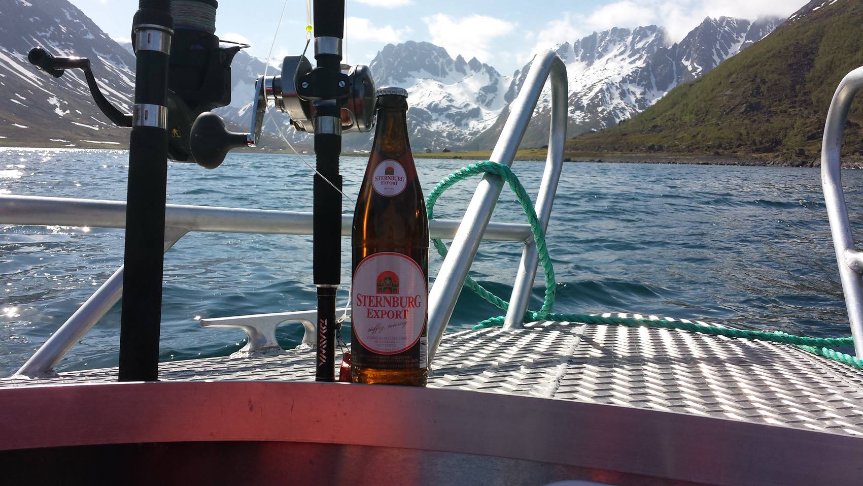 Eine Flasche Sternburg Export steht auf einem Boot vor einer Berglandschaft mit schneebedeckten Gipfeln und blauem Wasser.